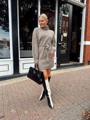 Woman wearing taupe turtle neck sweater dress with belt detail.