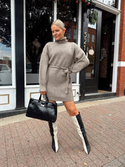 Woman wearing taupe turtle neck sweater dress with belt detail.