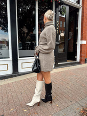 Woman wearing taupe turtle neck sweater dress with belt detail.