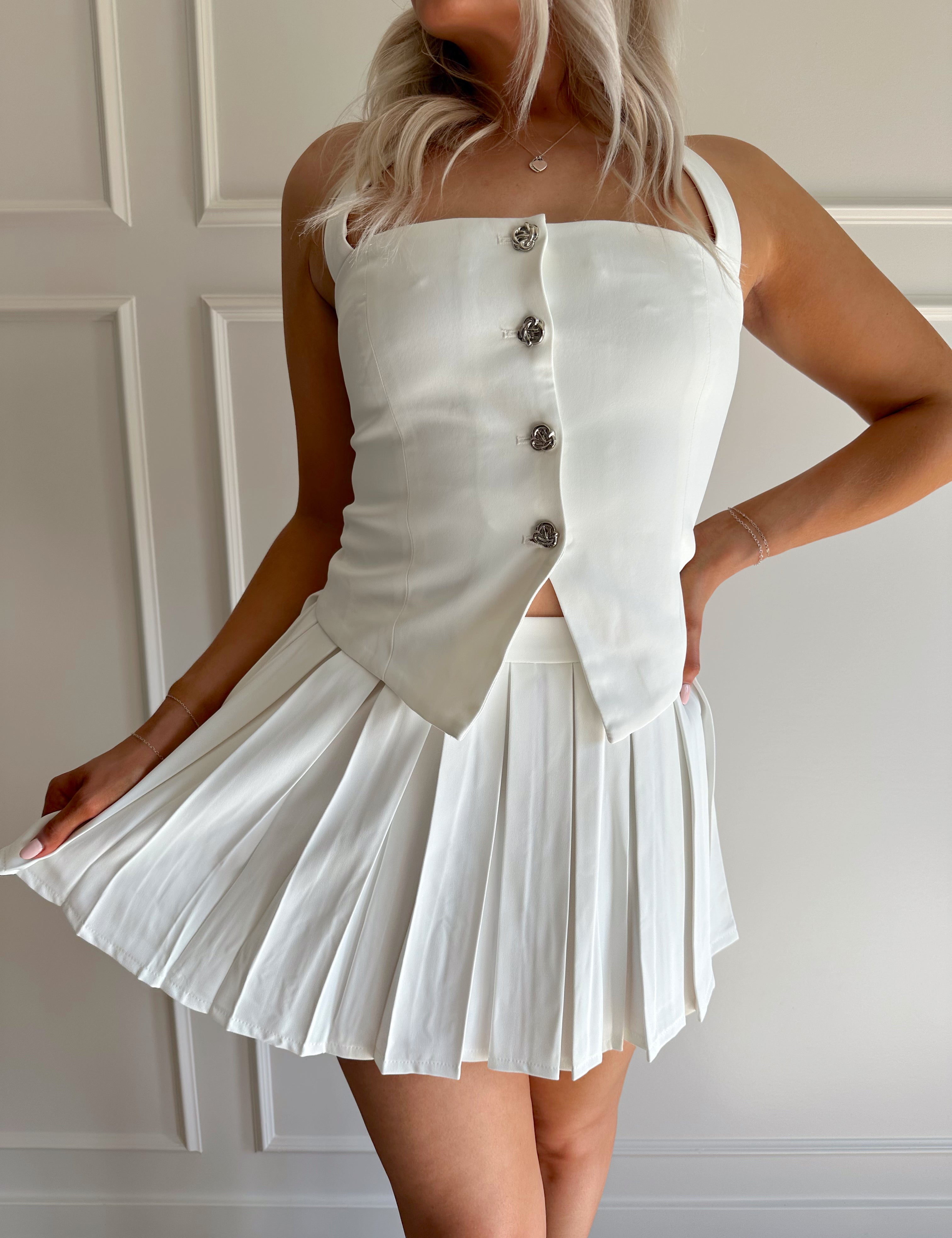 Woman wearing white two piece set with halter top with button detailing, and pleated mini skirt.
