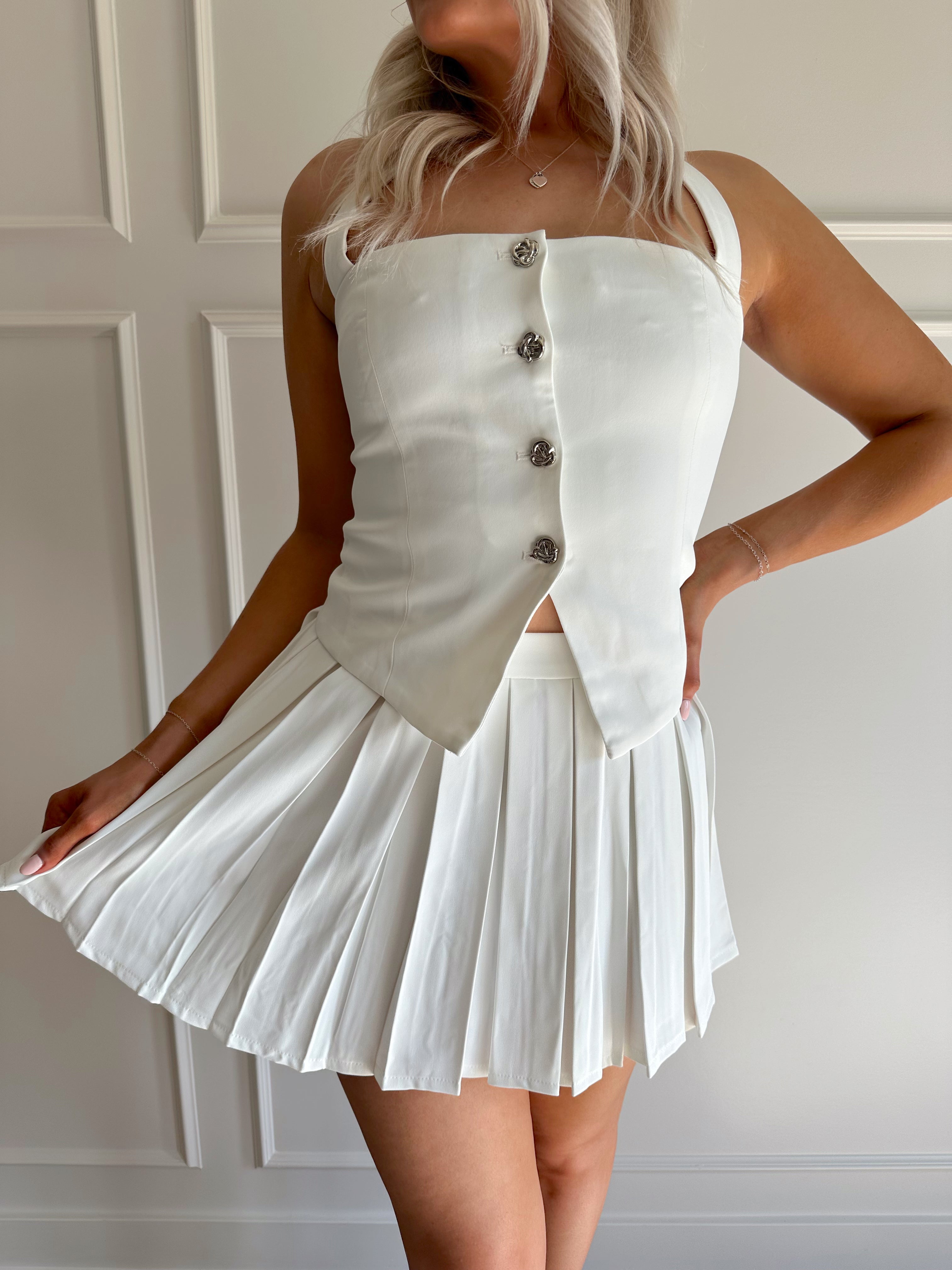 Woman wearing white two piece set with halter top with button detailing, and pleated mini skirt.