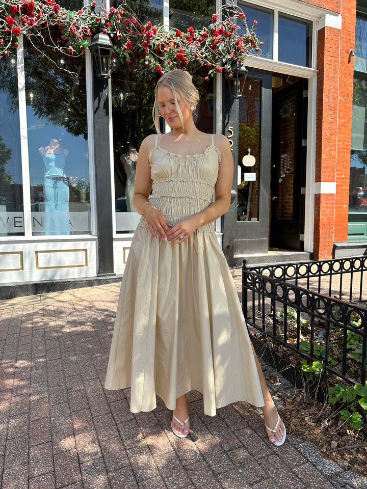 Woman wearing tan maxi dress with smocked bodice, adjustable spaghetti straps, and side slit. 