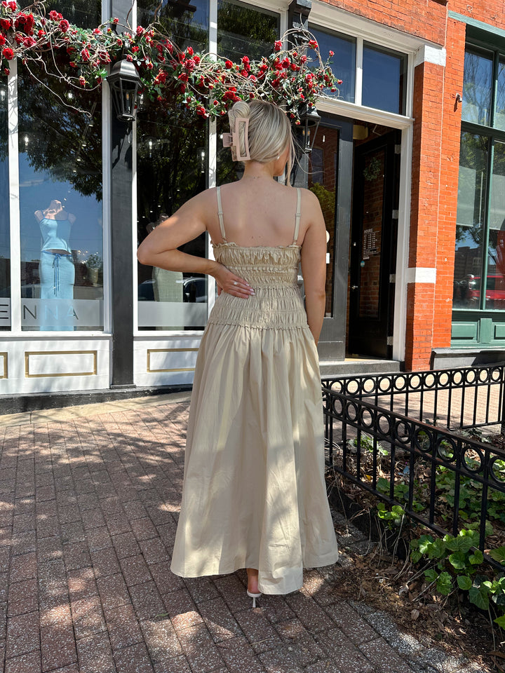 Woman wearing tan maxi dress with smocked bodice, adjustable spaghetti straps, and side slit. 