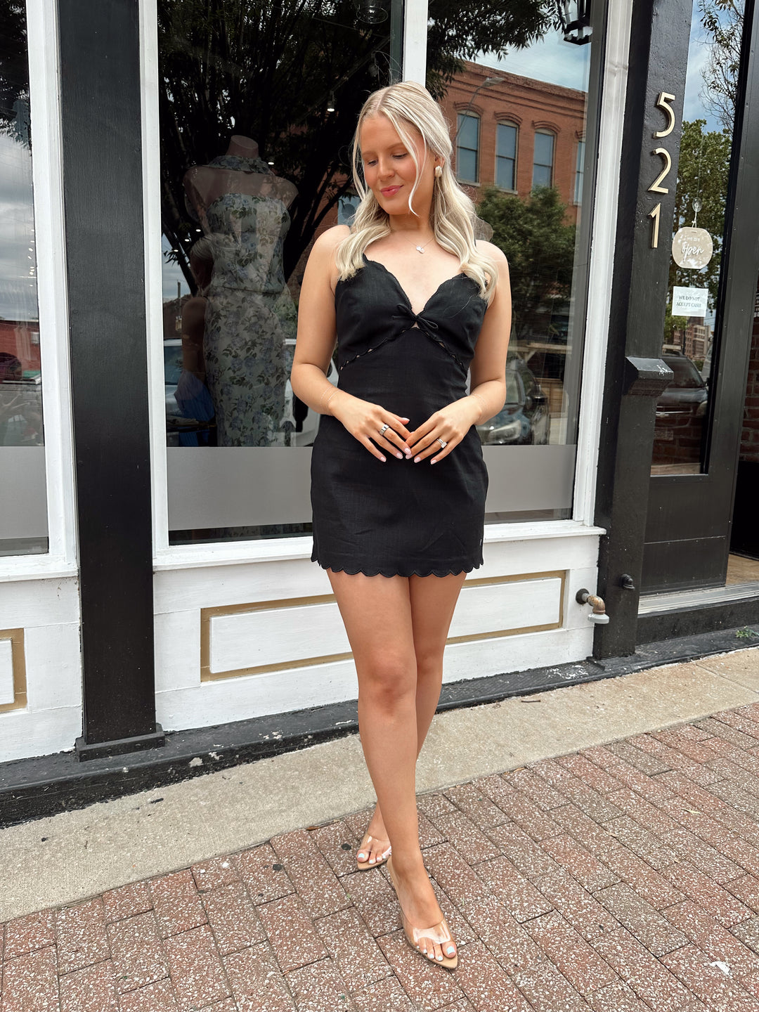 Woman wearing black linen mini dress with v-neckline and scallop hem.