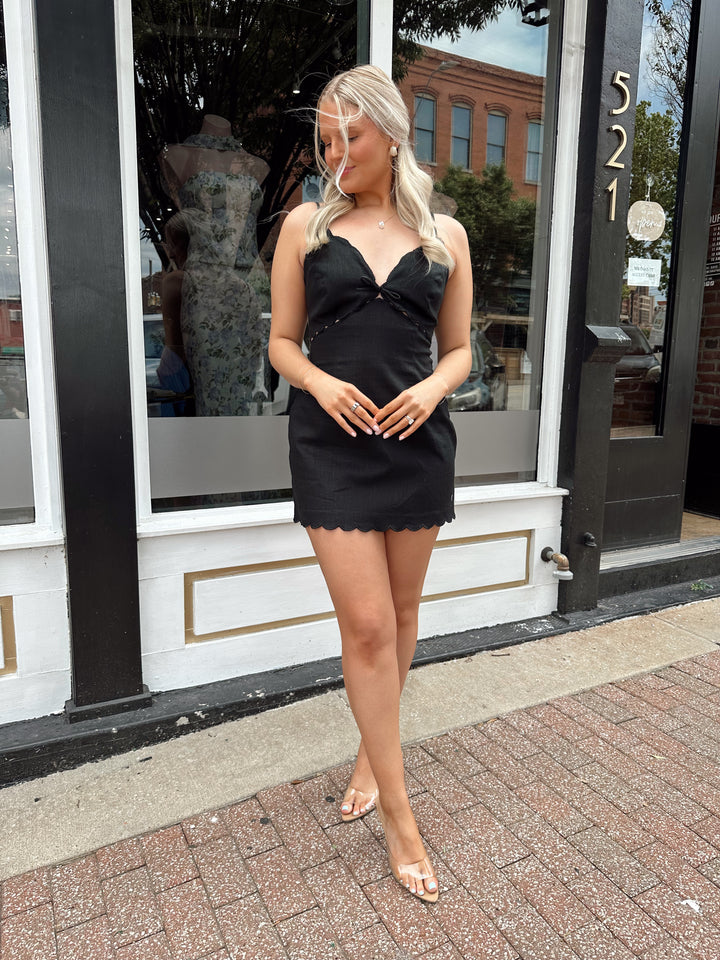 Woman wearing black linen mini dress with v-neckline and scallop hem.