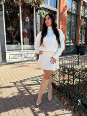 Woman wearing cream colored sweater set with pleated mini skirt. 
