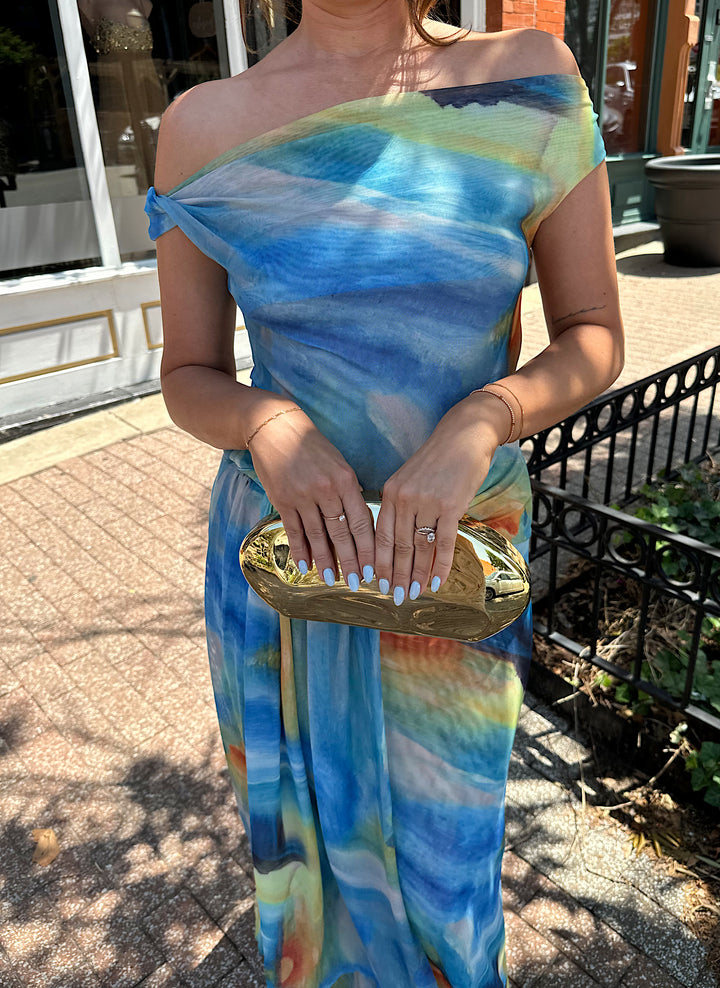Woman wearing off the shoulder, blue mesh maxi dress with watercolor detailing, and draping sash. 