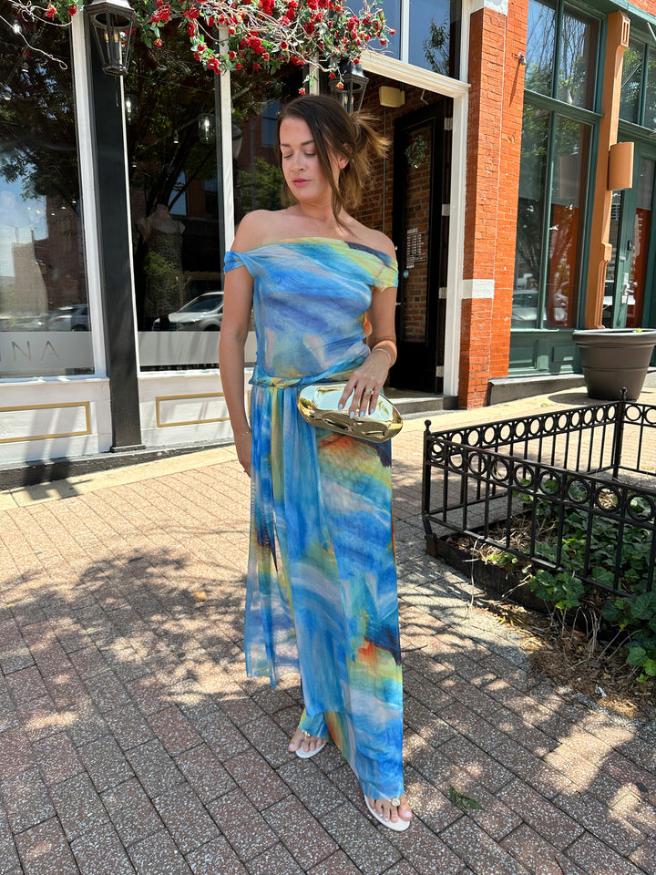 Woman wearing off the shoulder, blue mesh maxi dress with watercolor detailing, and draping sash. 