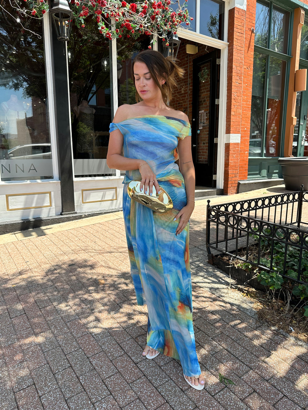 Woman wearing off the shoulder, blue mesh maxi dress with watercolor detailing, and draping sash. 