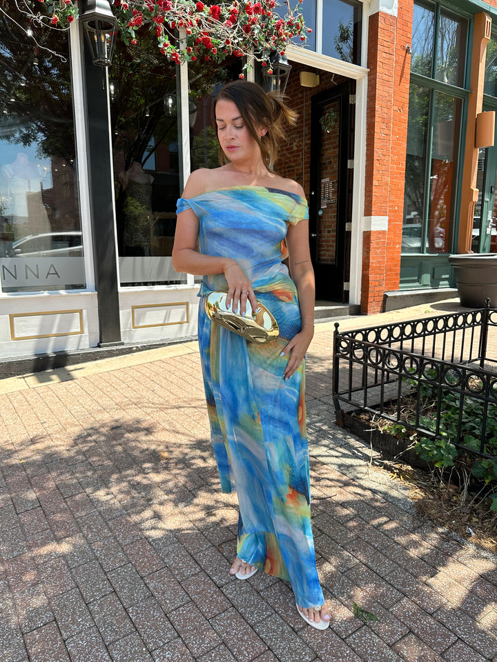 Woman wearing off the shoulder, blue mesh maxi dress with watercolor detailing, and draping sash. 