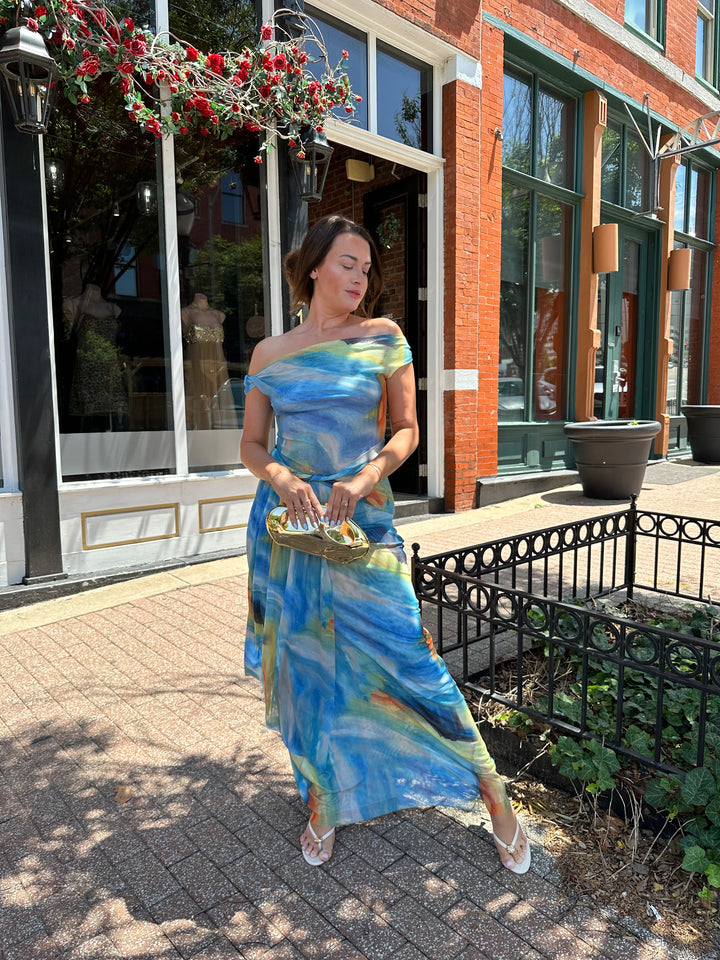 Woman wearing off the shoulder, blue mesh maxi dress with watercolor detailing, and draping sash. 