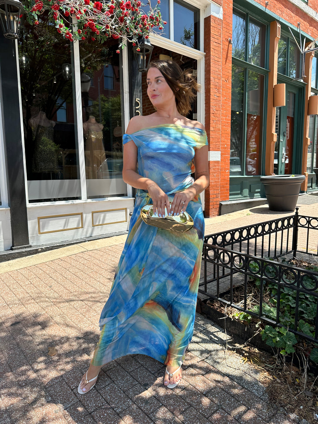 Woman wearing off the shoulder, blue mesh maxi dress with watercolor detailing, and draping sash. 