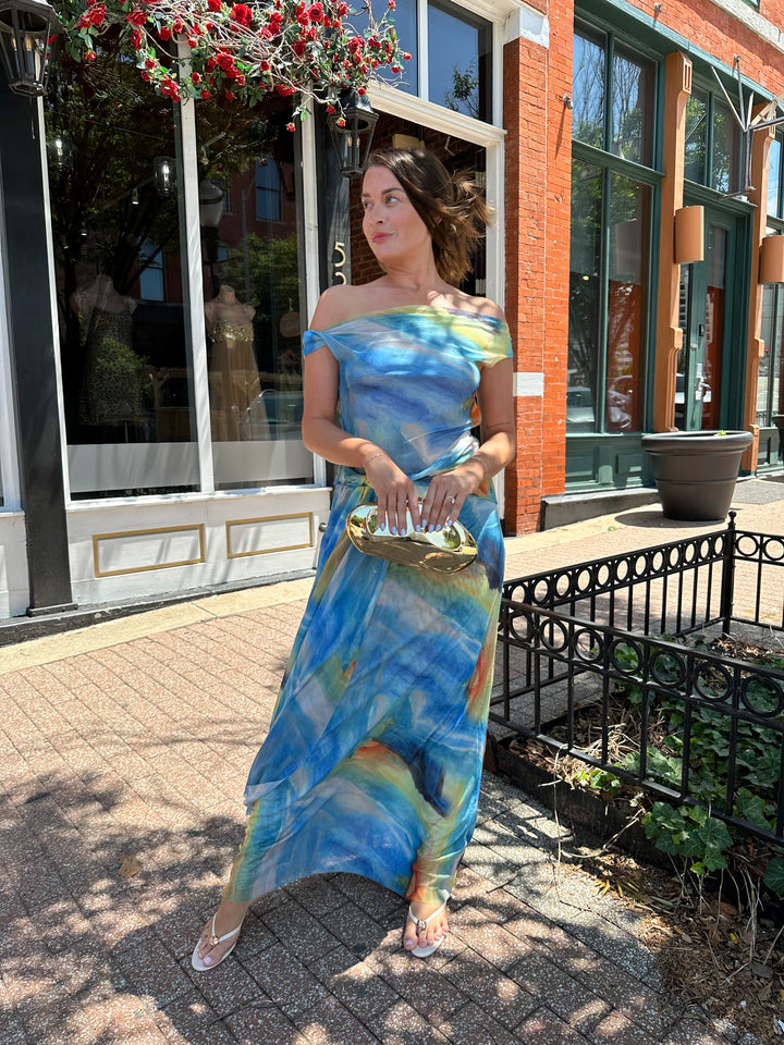 Woman wearing off the shoulder, blue mesh maxi dress with watercolor detailing, and draping sash. 