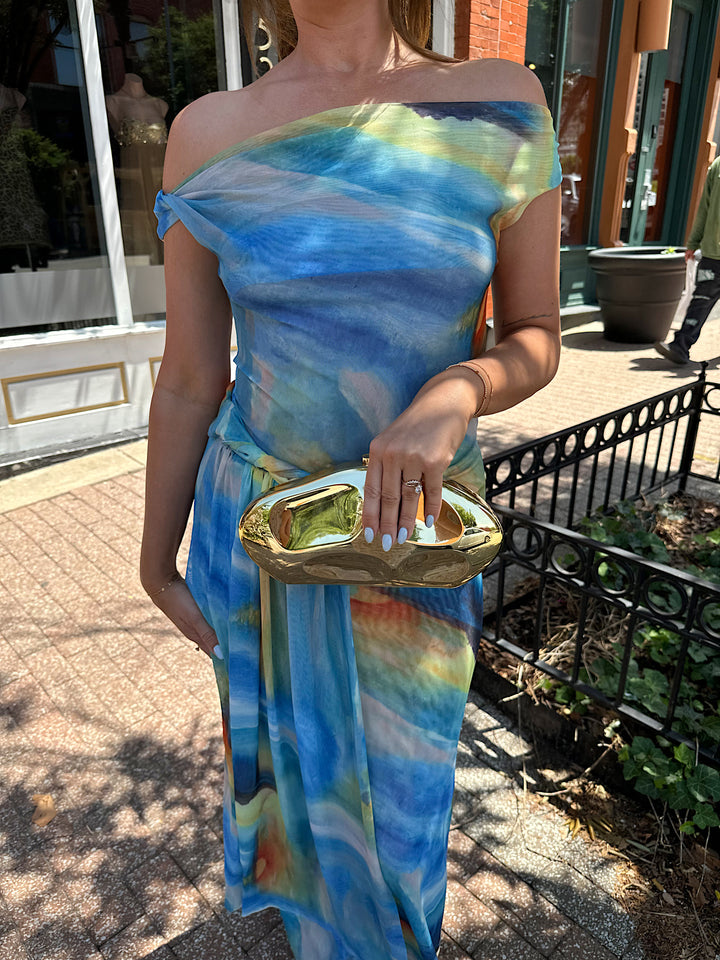 Woman wearing off the shoulder, blue mesh maxi dress with watercolor detailing, and draping sash. 