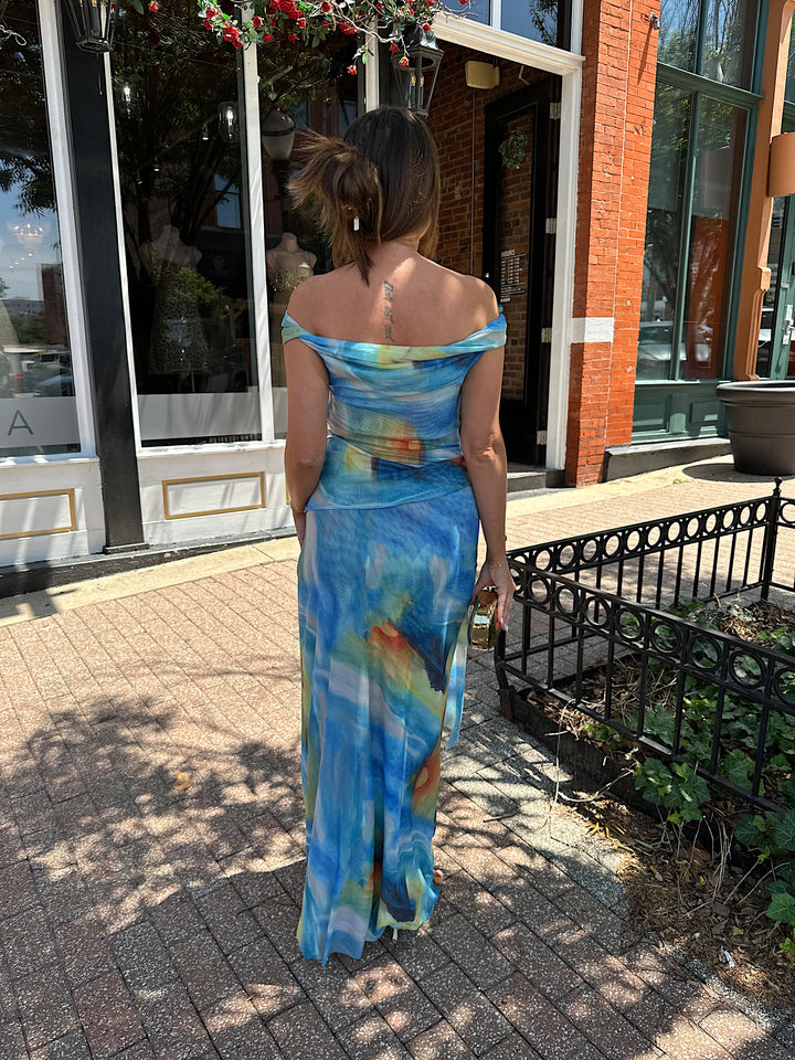 Woman wearing off the shoulder, blue mesh maxi dress with watercolor detailing, and draping sash. 