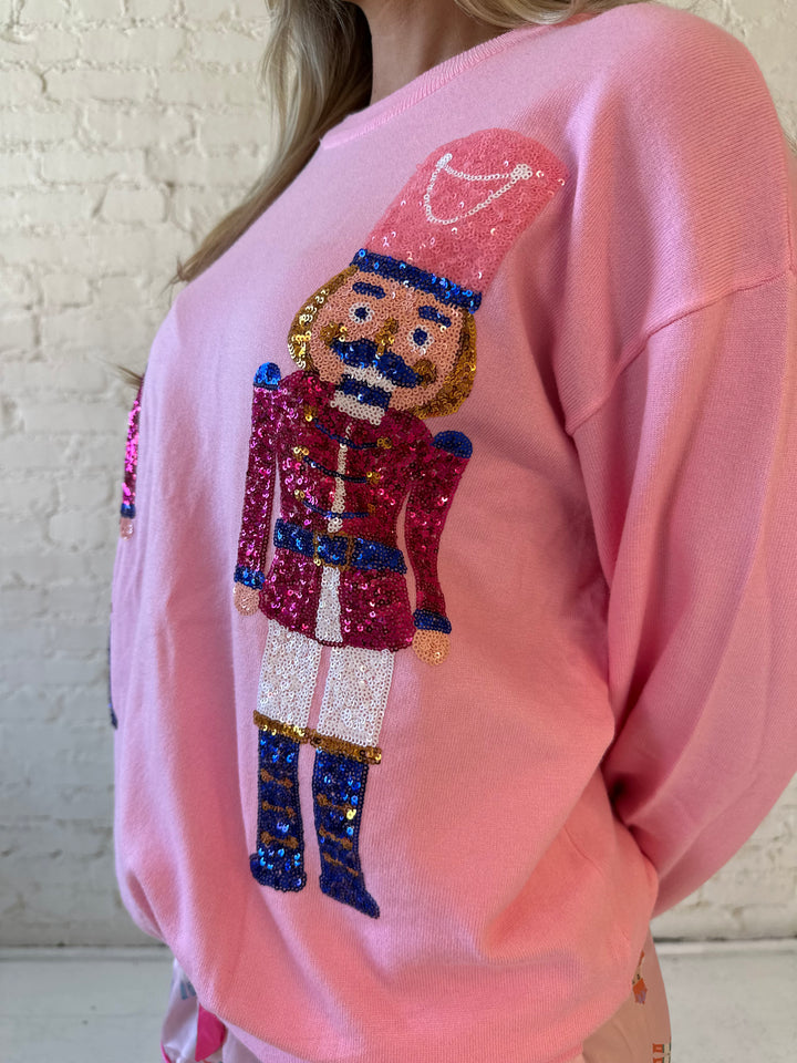 Woman wearing blush pink knit crewneck sweater with oversized sequin nutcrackers.