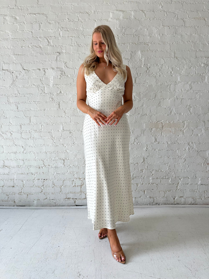 Woman wearing cream polka dot satin maxi dress with v-neckline and open back with tie detail. 