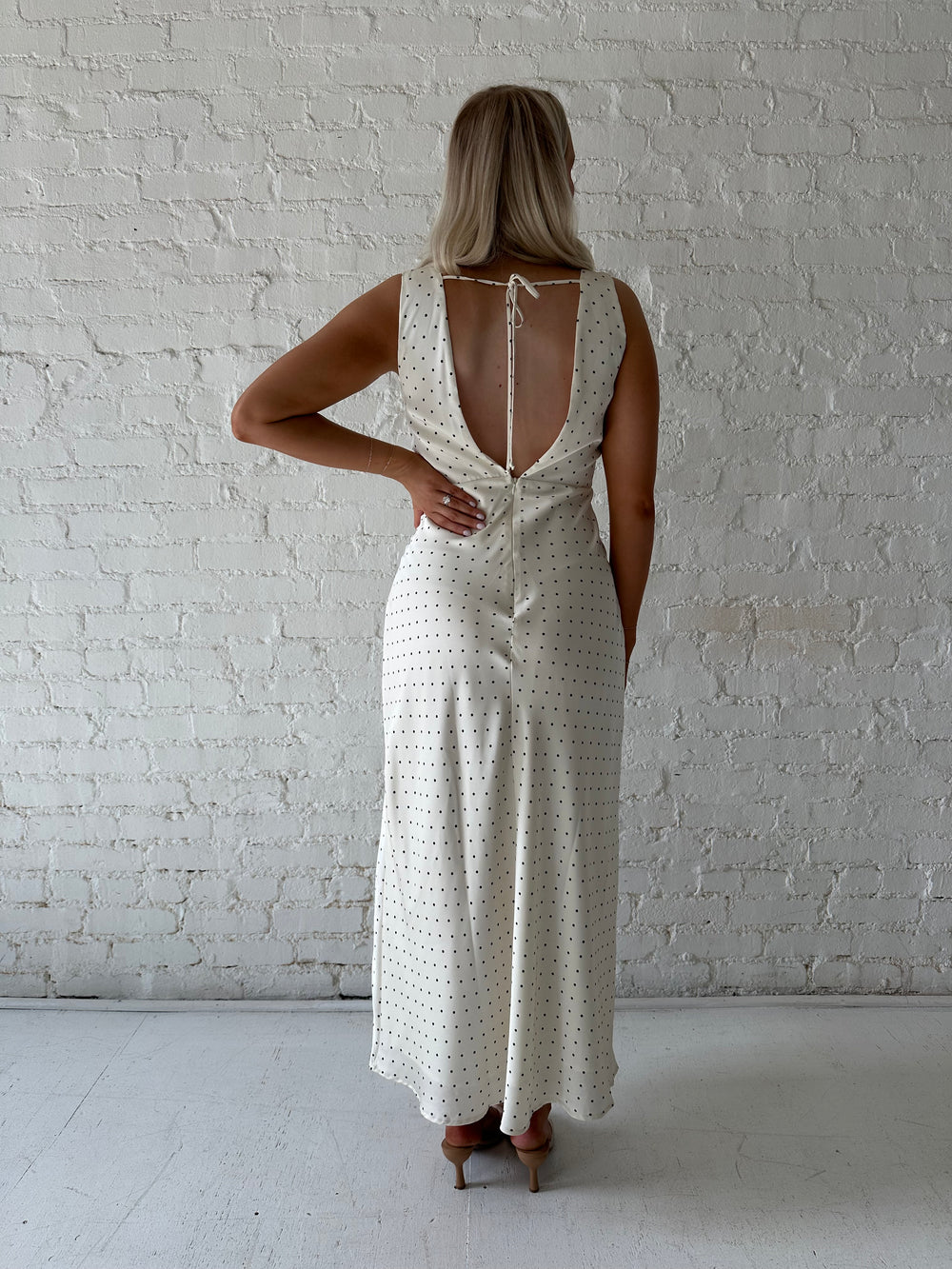 Woman wearing cream polka dot satin maxi dress with v-neckline and open back with tie detail. 