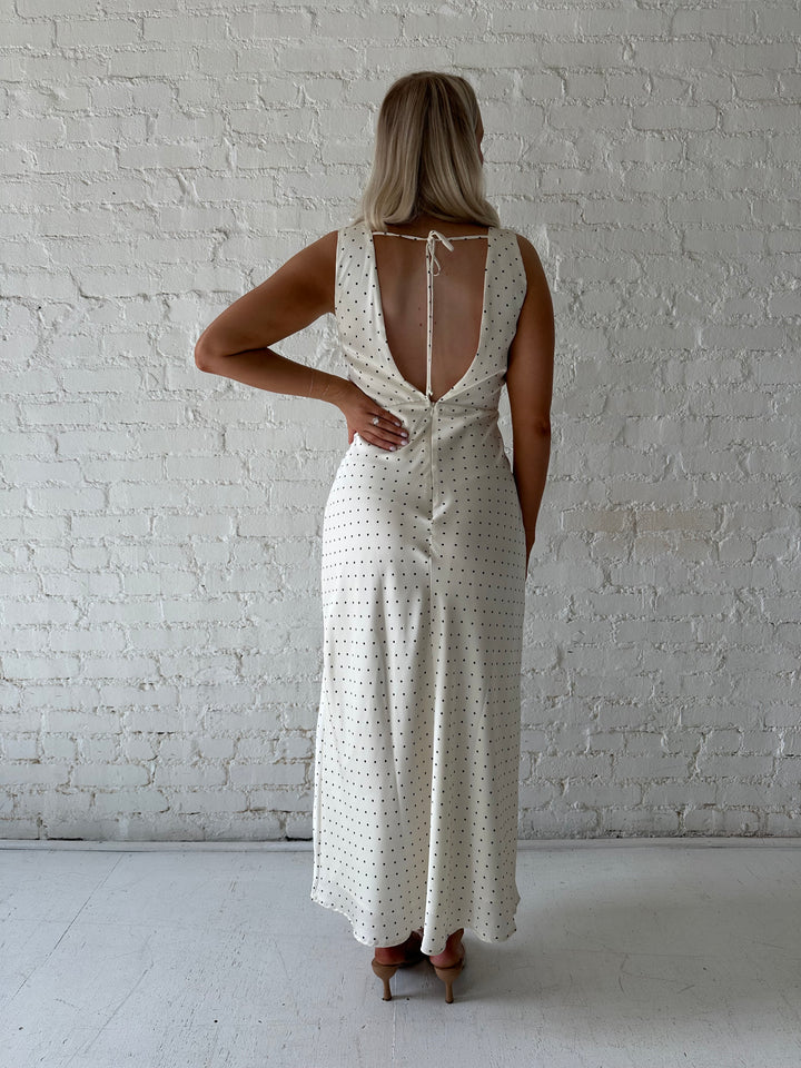 Woman wearing cream polka dot satin maxi dress with v-neckline and open back with tie detail. 