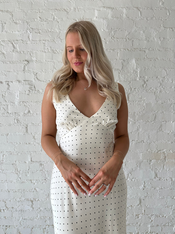 Woman wearing cream polka dot satin maxi dress with v-neckline and open back with tie detail. 