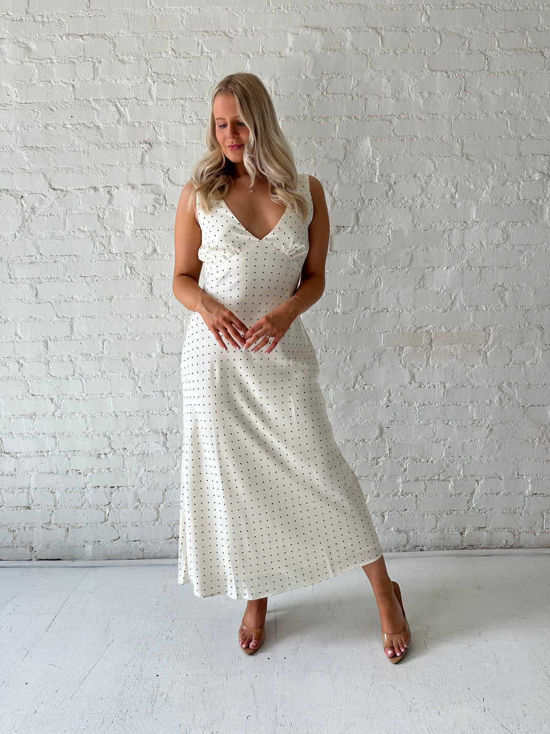 Woman wearing cream polka dot satin maxi dress with v-neckline and open back with tie detail. 