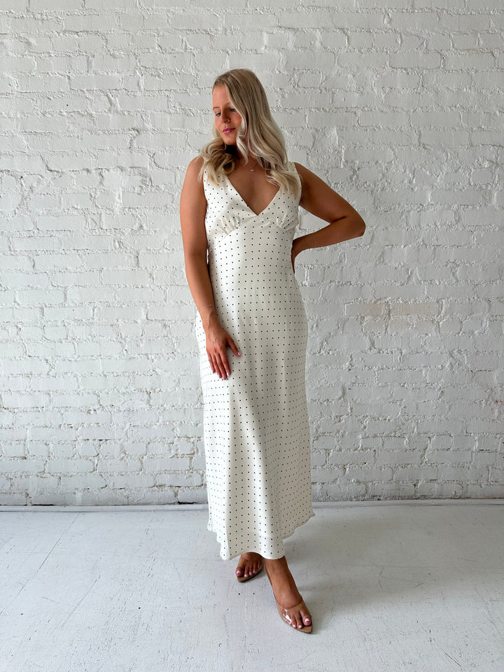 Woman wearing cream polka dot satin maxi dress with v-neckline and open back with tie detail. 