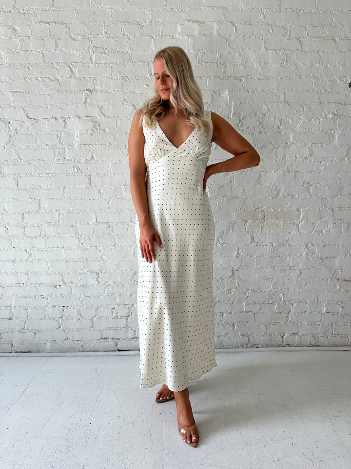 Woman wearing cream polka dot satin maxi dress with v-neckline and open back with tie detail. 