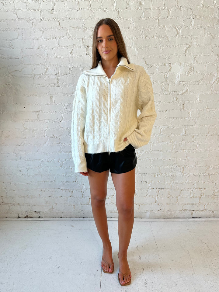 Woman wearing cream fuzzy cable knit zip up jacket with black leather shorts. 