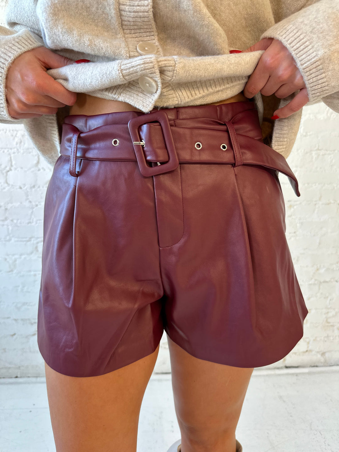 Woman wearing burgundy faux leather high waisted belted shorts with beige knit button down cardigan. 