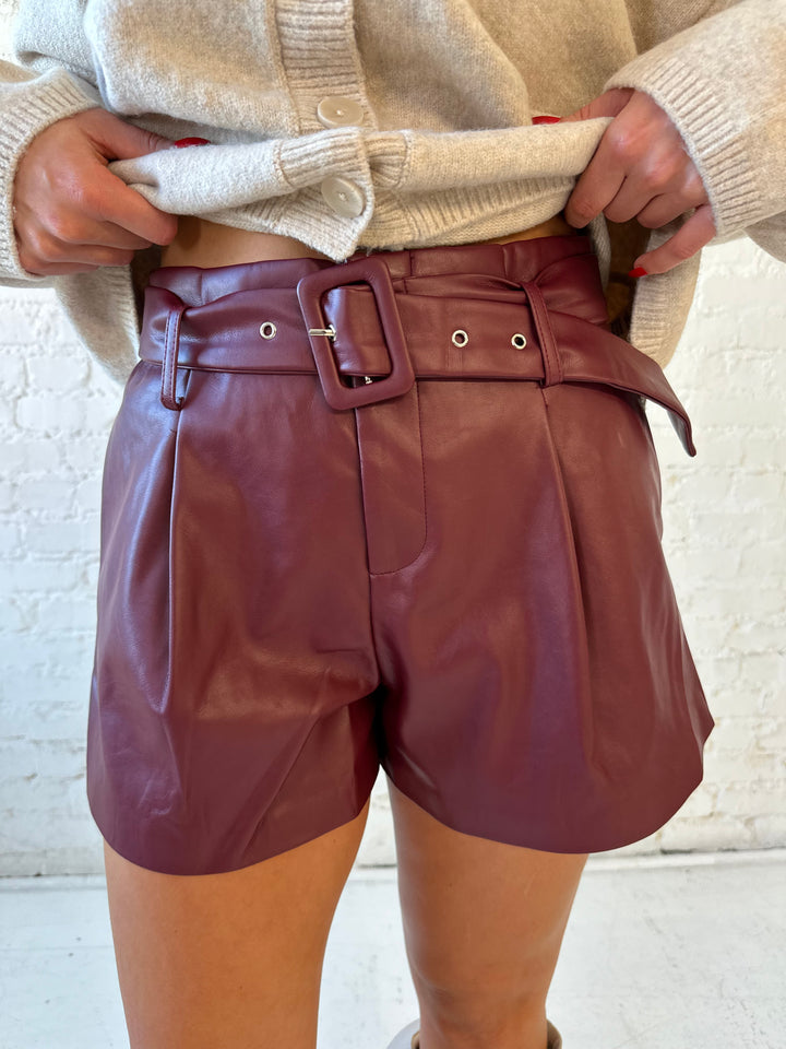 Woman wearing burgundy faux leather high waisted belted shorts with beige knit button down cardigan. 
