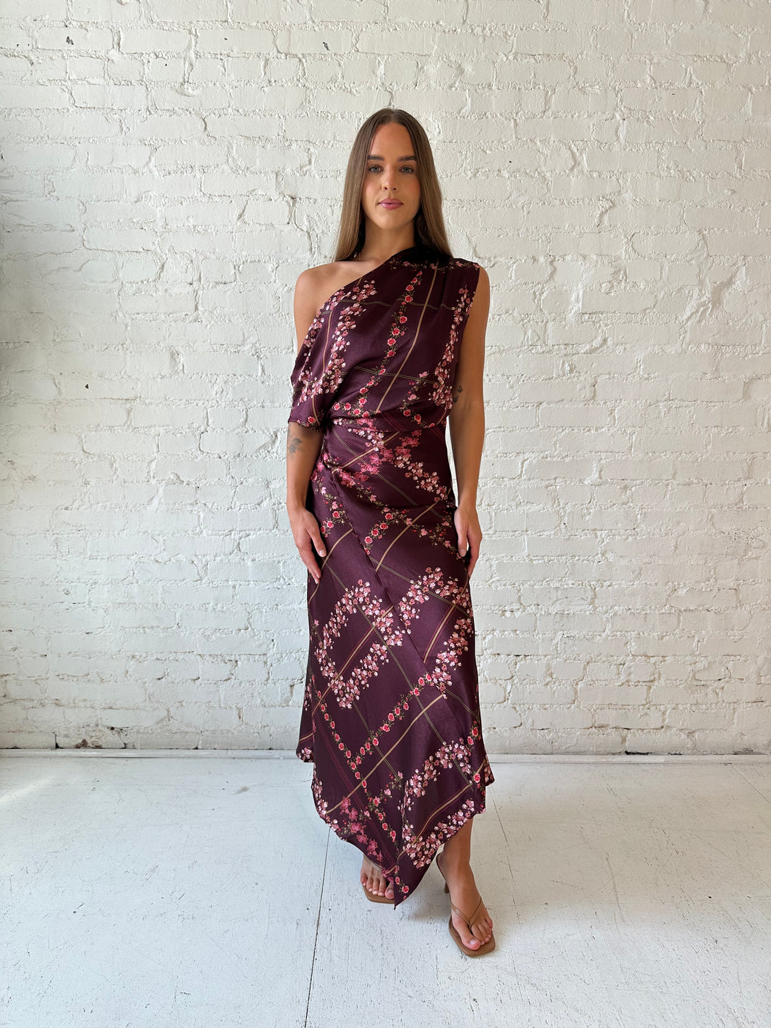 Woman wearing burgundy floral satin maxi dress with asymmetrical neckline, and flutter hem.