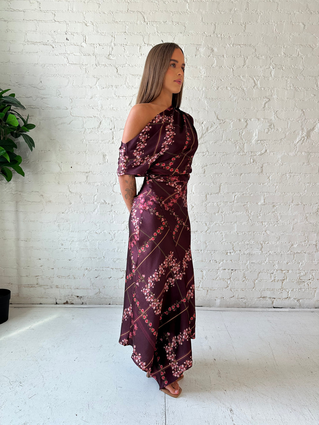 Woman wearing burgundy floral satin maxi dress with asymmetrical neckline, and flutter hem.
