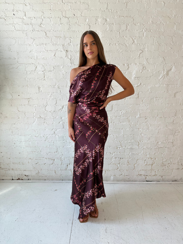 Woman wearing burgundy floral satin maxi dress with asymmetrical neckline, and flutter hem.
