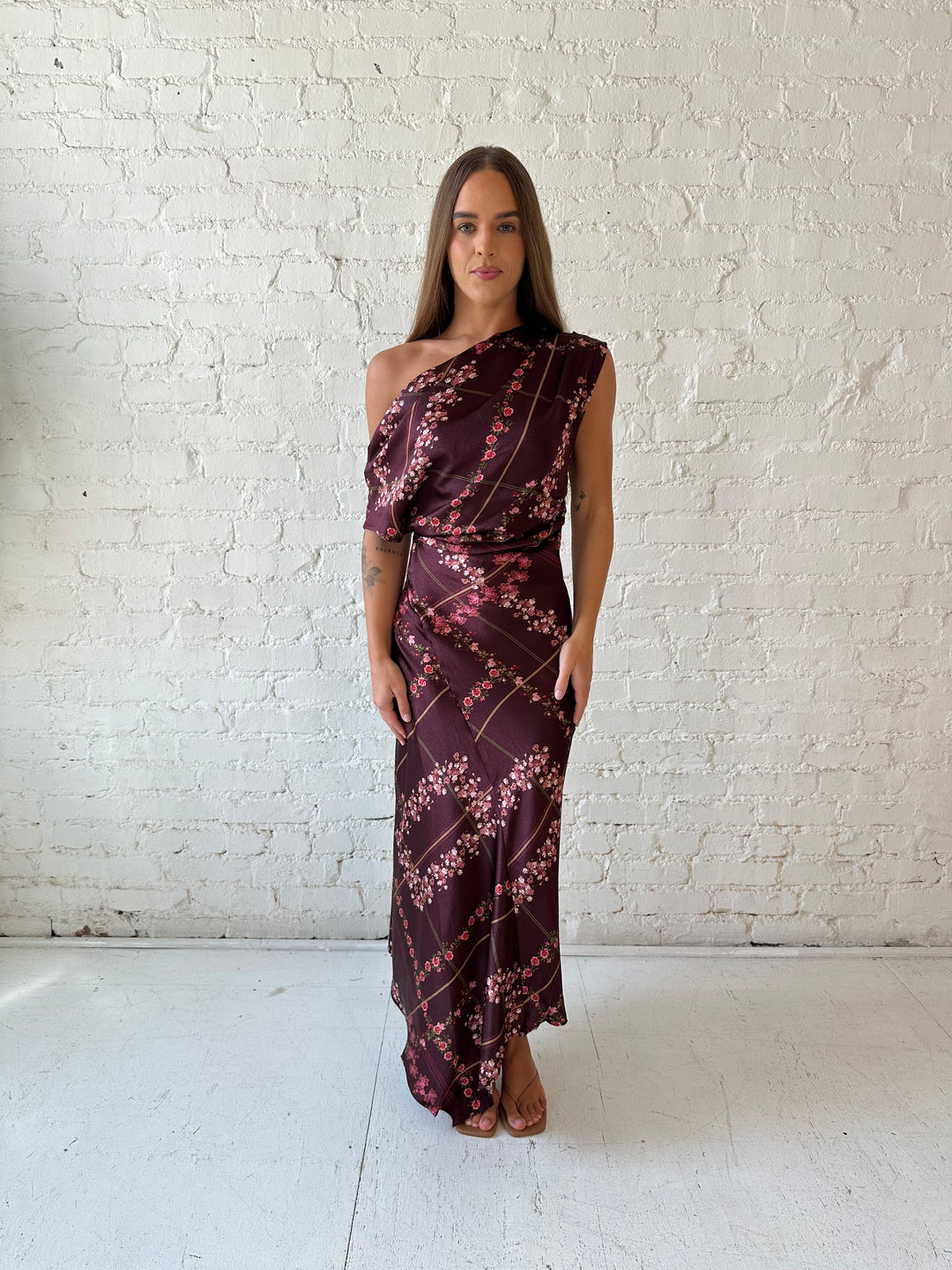 Woman wearing burgundy floral satin maxi dress with asymmetrical neckline, and flutter hem.