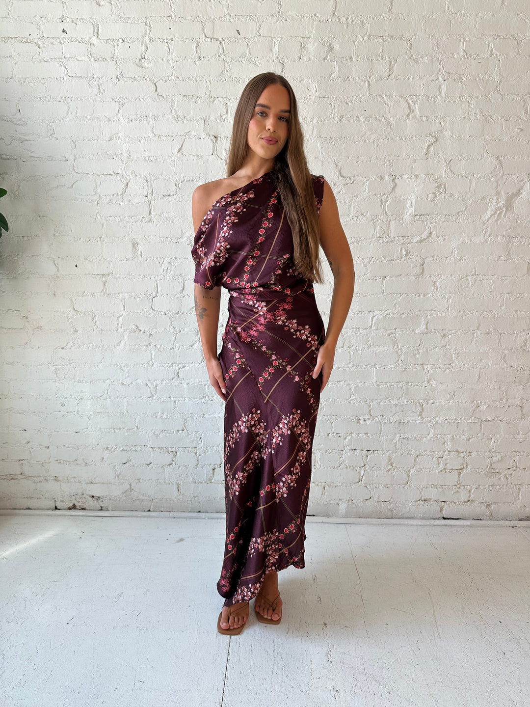 Woman wearing burgundy floral satin maxi dress with asymmetrical neckline, and flutter hem.