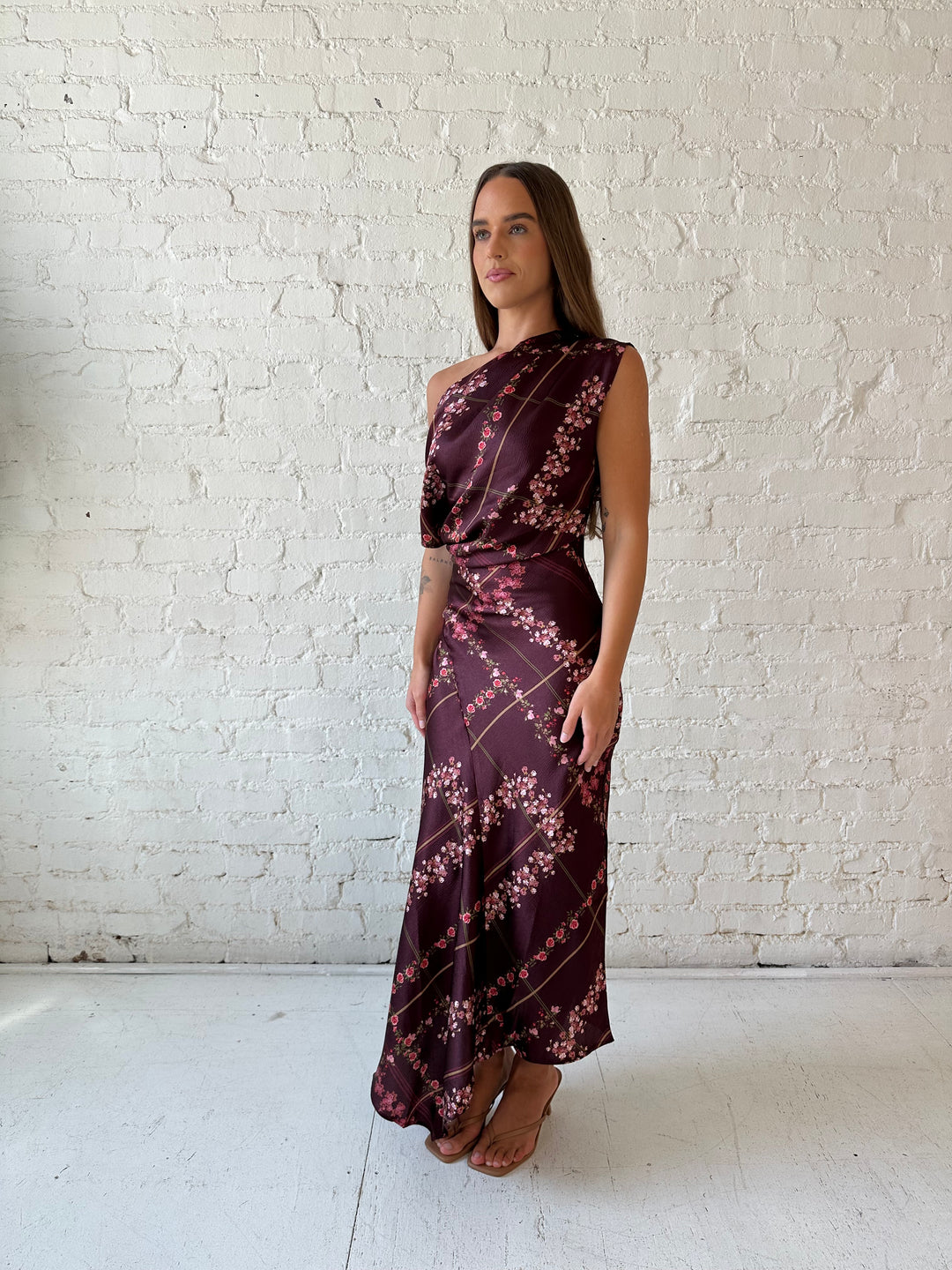 Woman wearing burgundy floral satin maxi dress with asymmetrical neckline, and flutter hem.