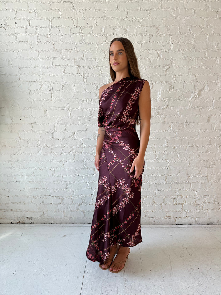 Woman wearing burgundy floral satin maxi dress with asymmetrical neckline, and flutter hem.
