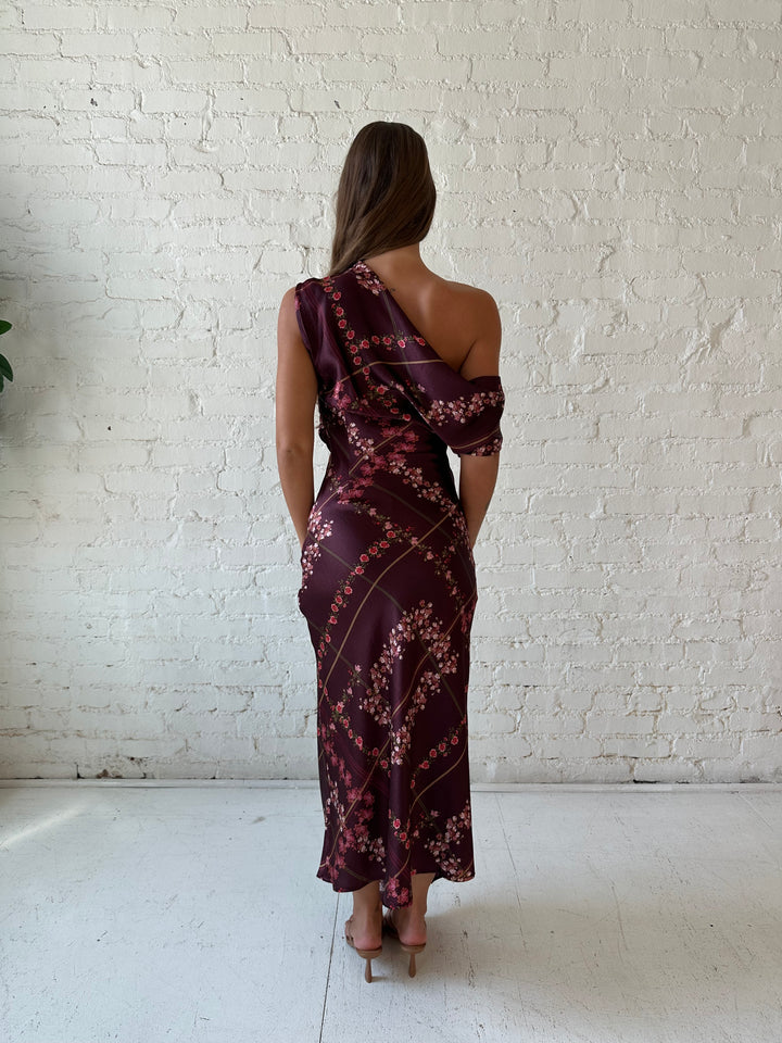 Woman wearing burgundy floral satin maxi dress with asymmetrical neckline, and flutter hem.