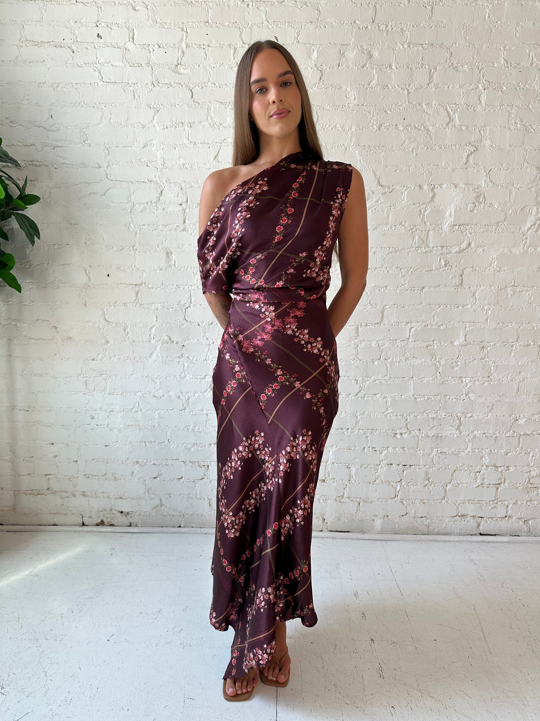 Woman wearing burgundy floral satin maxi dress with asymmetrical neckline, and flutter hem.
