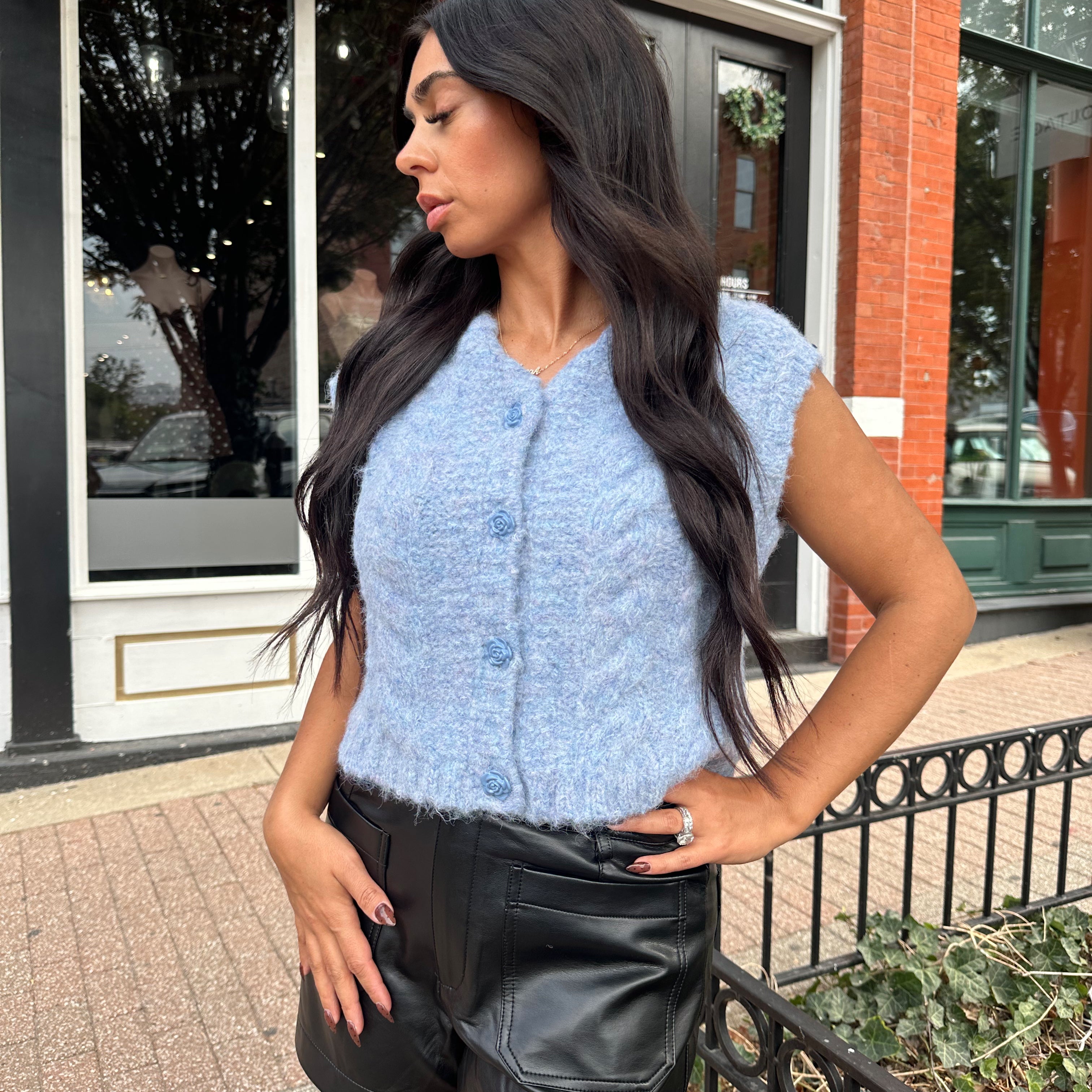 Woman wearing light blue knit sleeveless sweater vest with rose button detailing closure. 