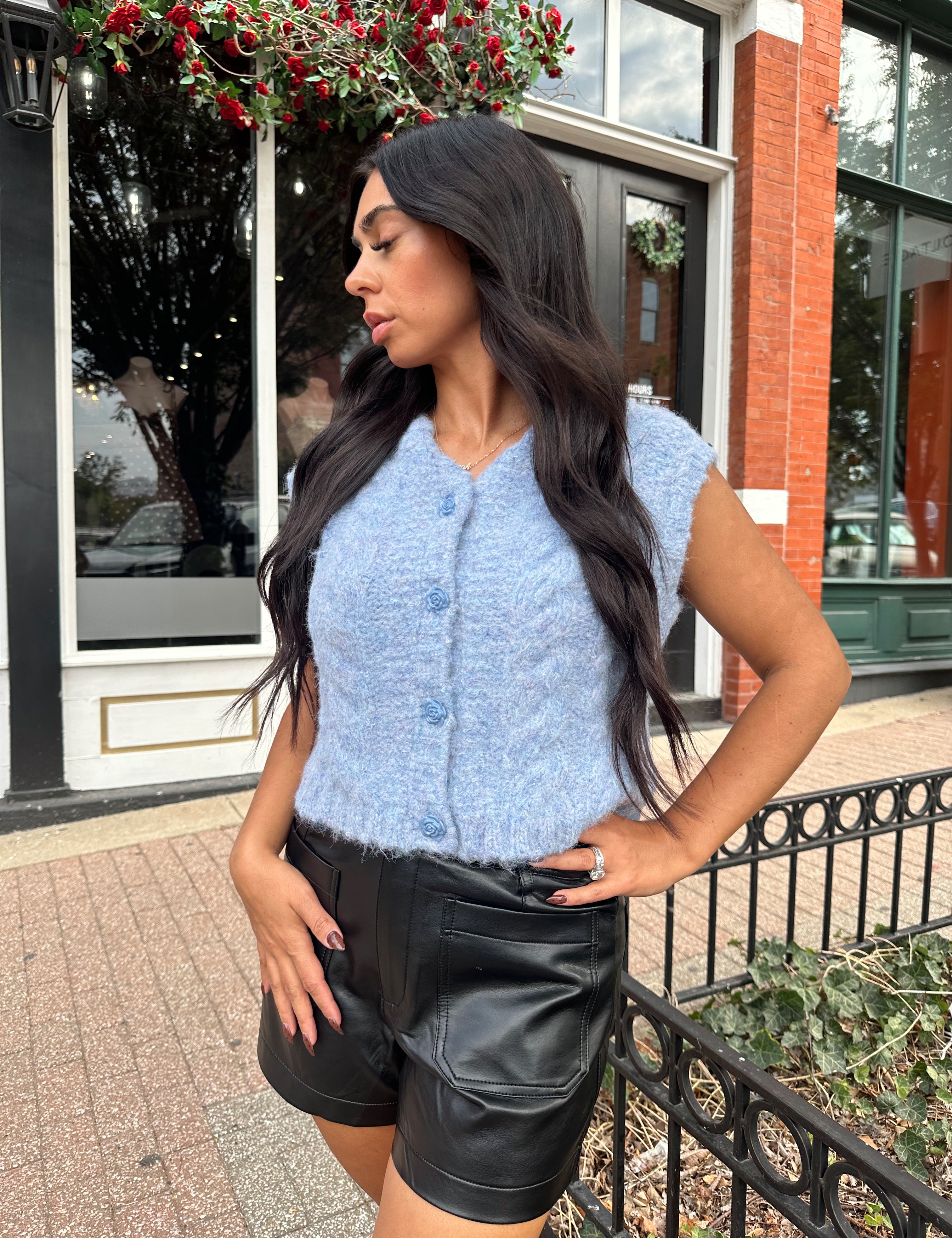 Woman wearing light blue knit sleeveless sweater vest with rose button detailing closure. 