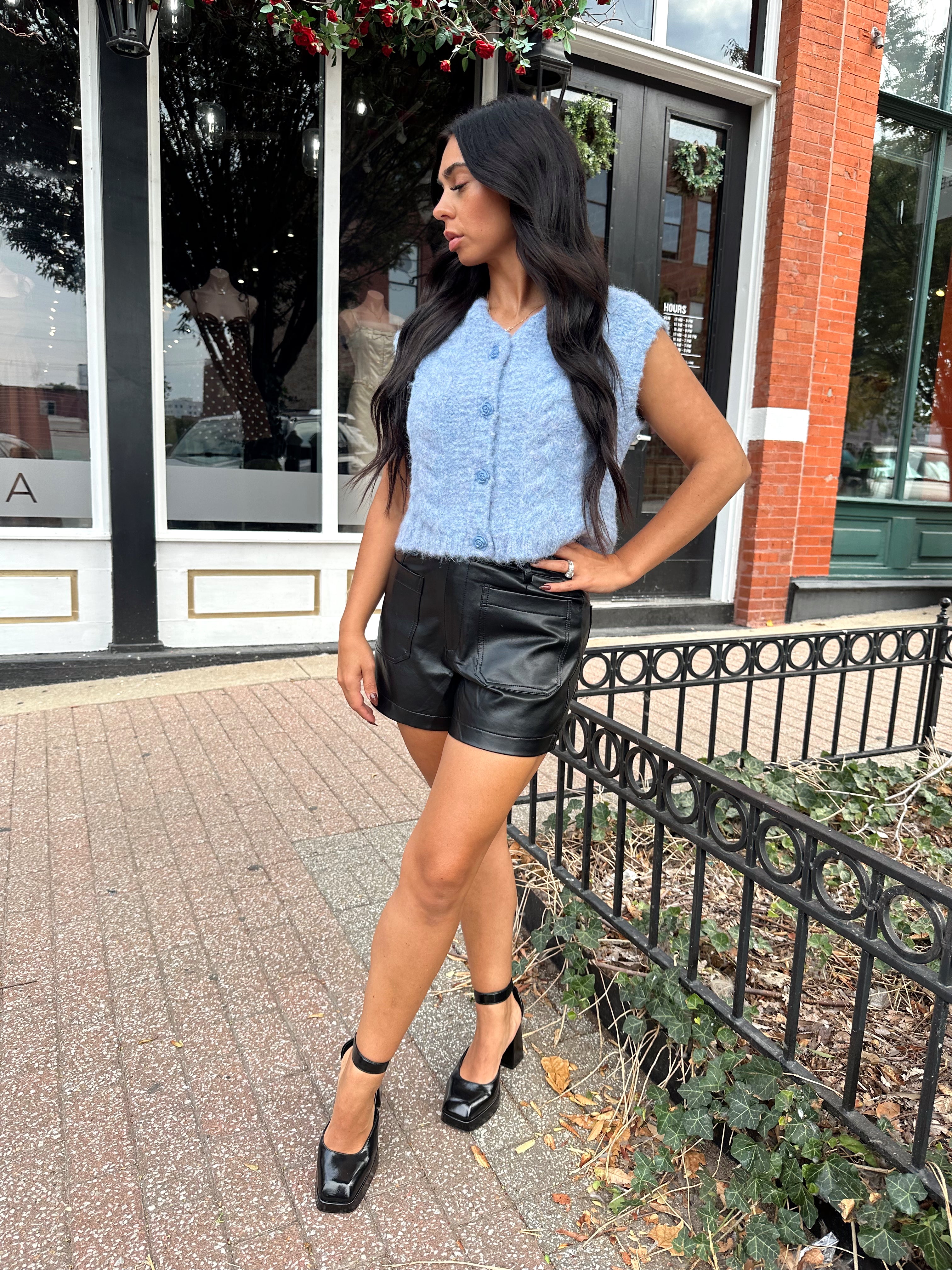 Woman wearing light blue knit sleeveless sweater vest with rose button detailing closure. 