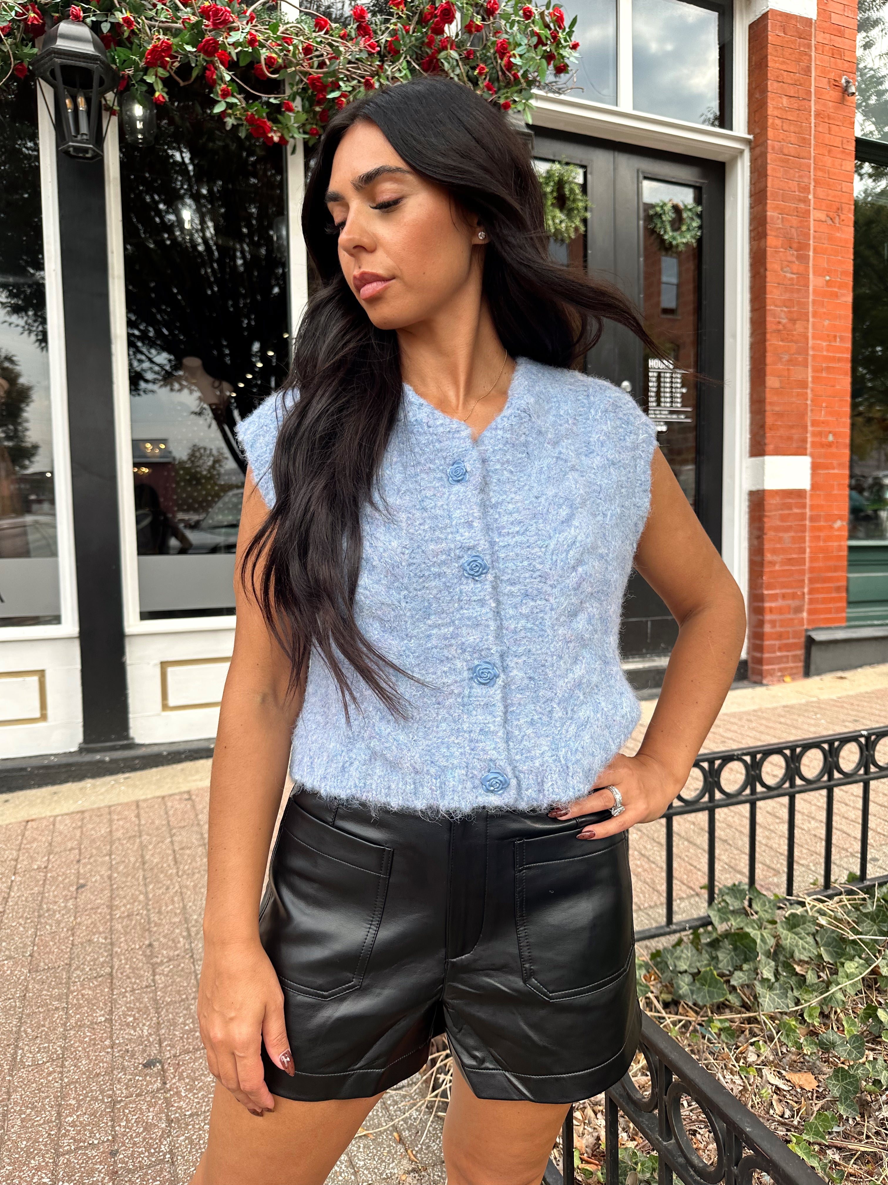 Woman wearing light blue knit sleeveless sweater vest with rose button detailing closure. 