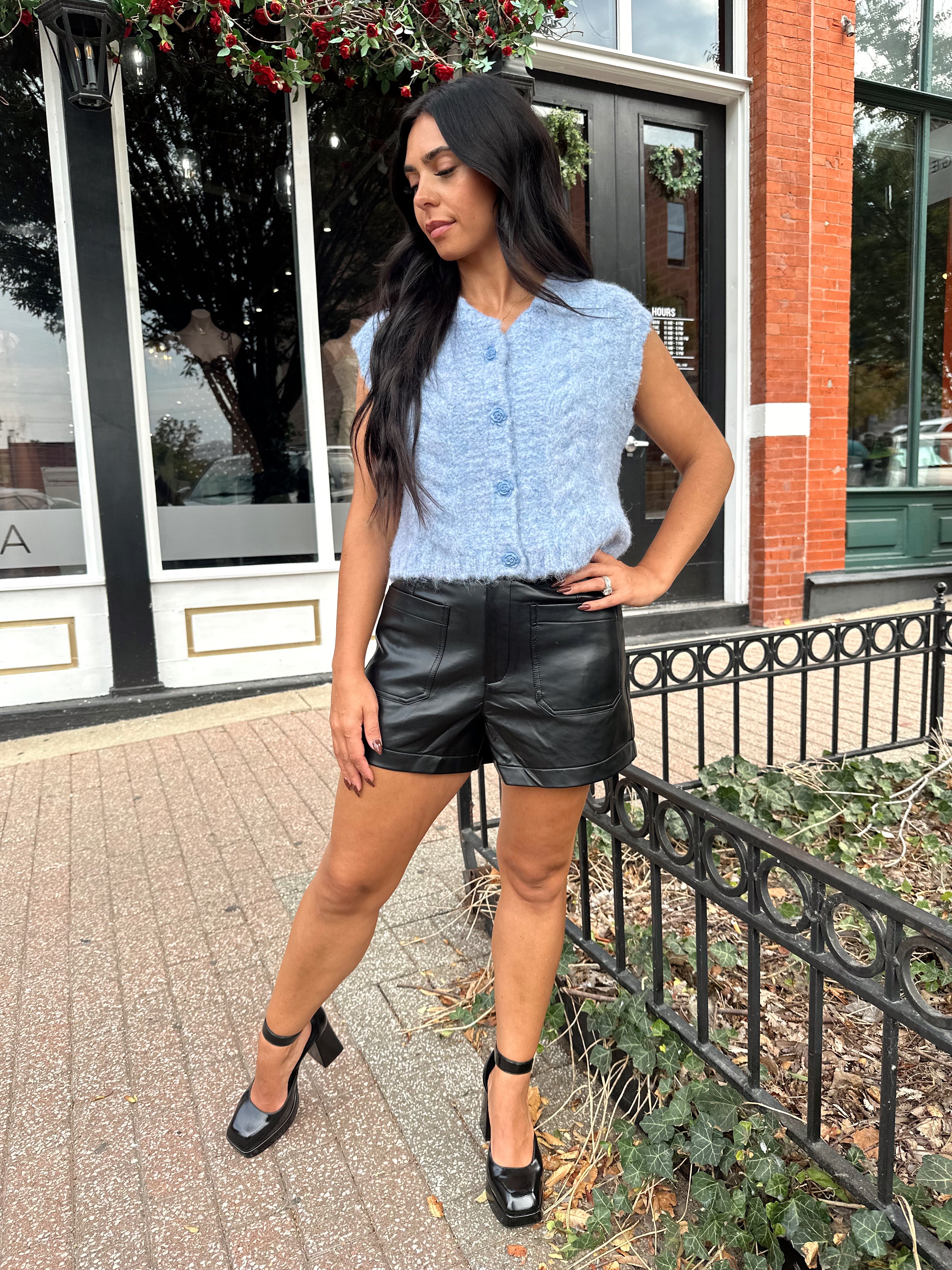 Woman wearing light blue knit sleeveless sweater vest with rose button detailing closure. 