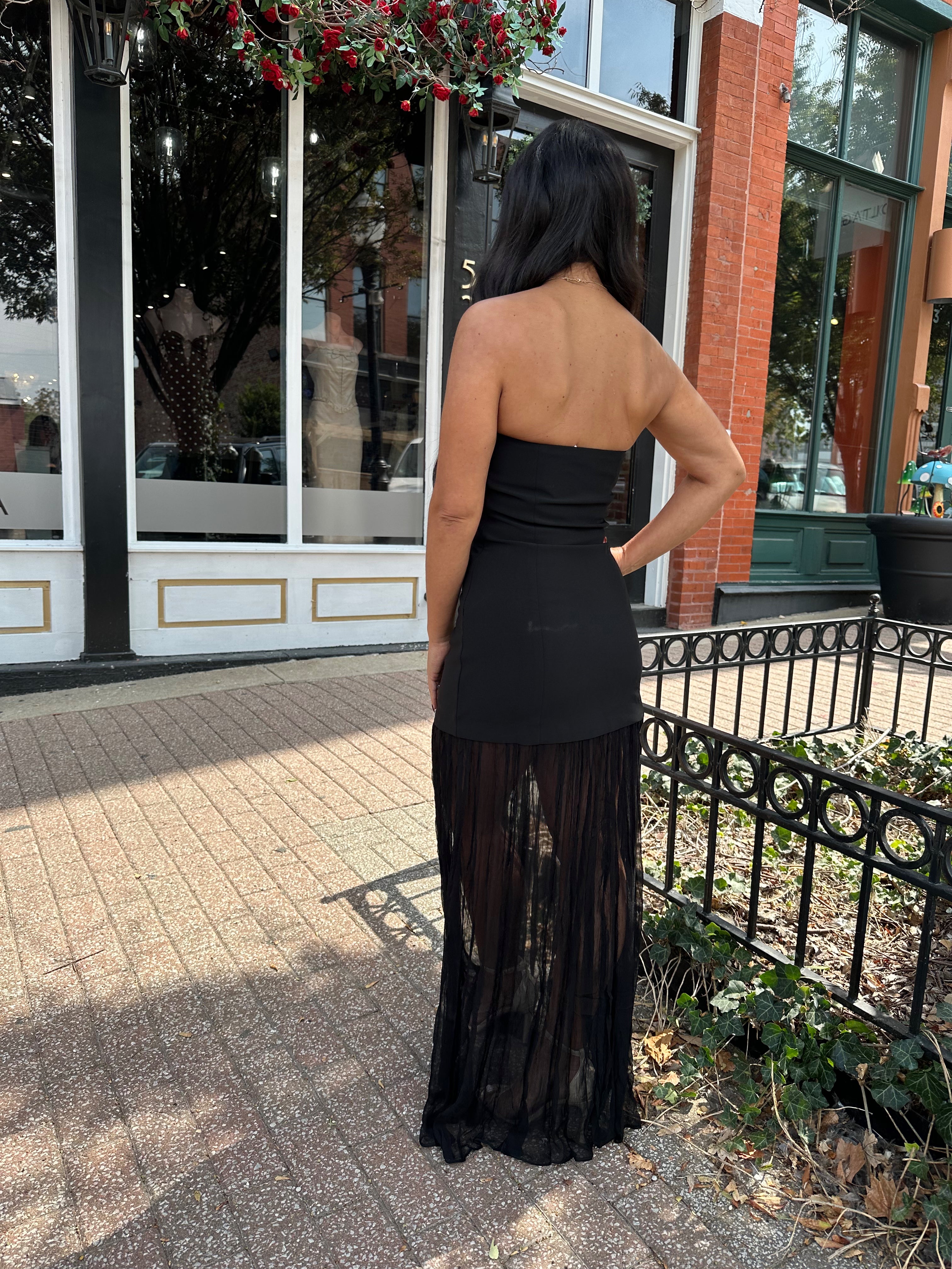 Woman wearing black strapless maxi dress with sheer overlay bottom.