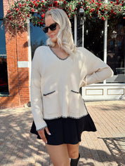 Woman wearing oatmeal sweater with black stitching and front pockets.