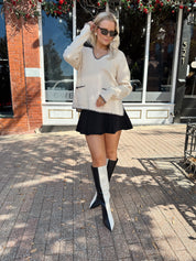Woman wearing oatmeal sweater with black stitching and front pockets.