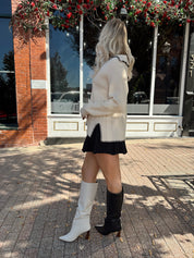 Woman wearing oatmeal sweater with black stitching and front pockets.