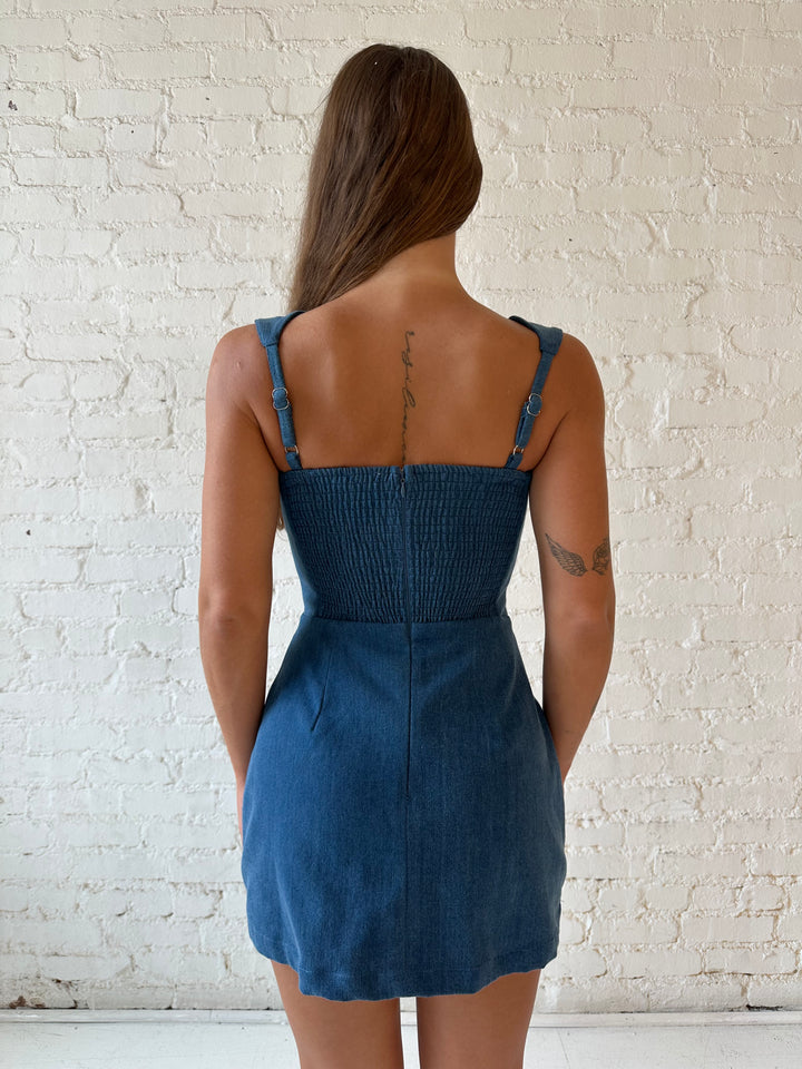 Woman wearing dark wash denim mini dress with square neckline, and built in shorts. 