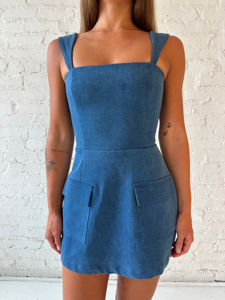 Woman wearing dark wash denim mini dress with square neckline, and built in shorts. 